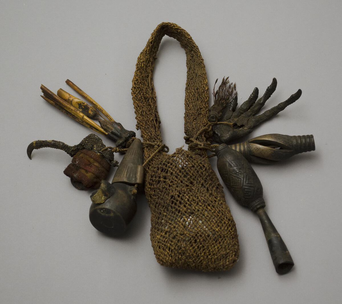 A bag made of vegetable fibers containing a wrapped bundle, with charms of wood, feathers, and animal feet attached