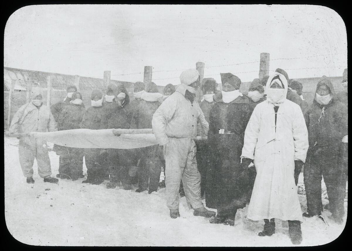 Men wearing masks carry a stretcher in front of a crowd of people