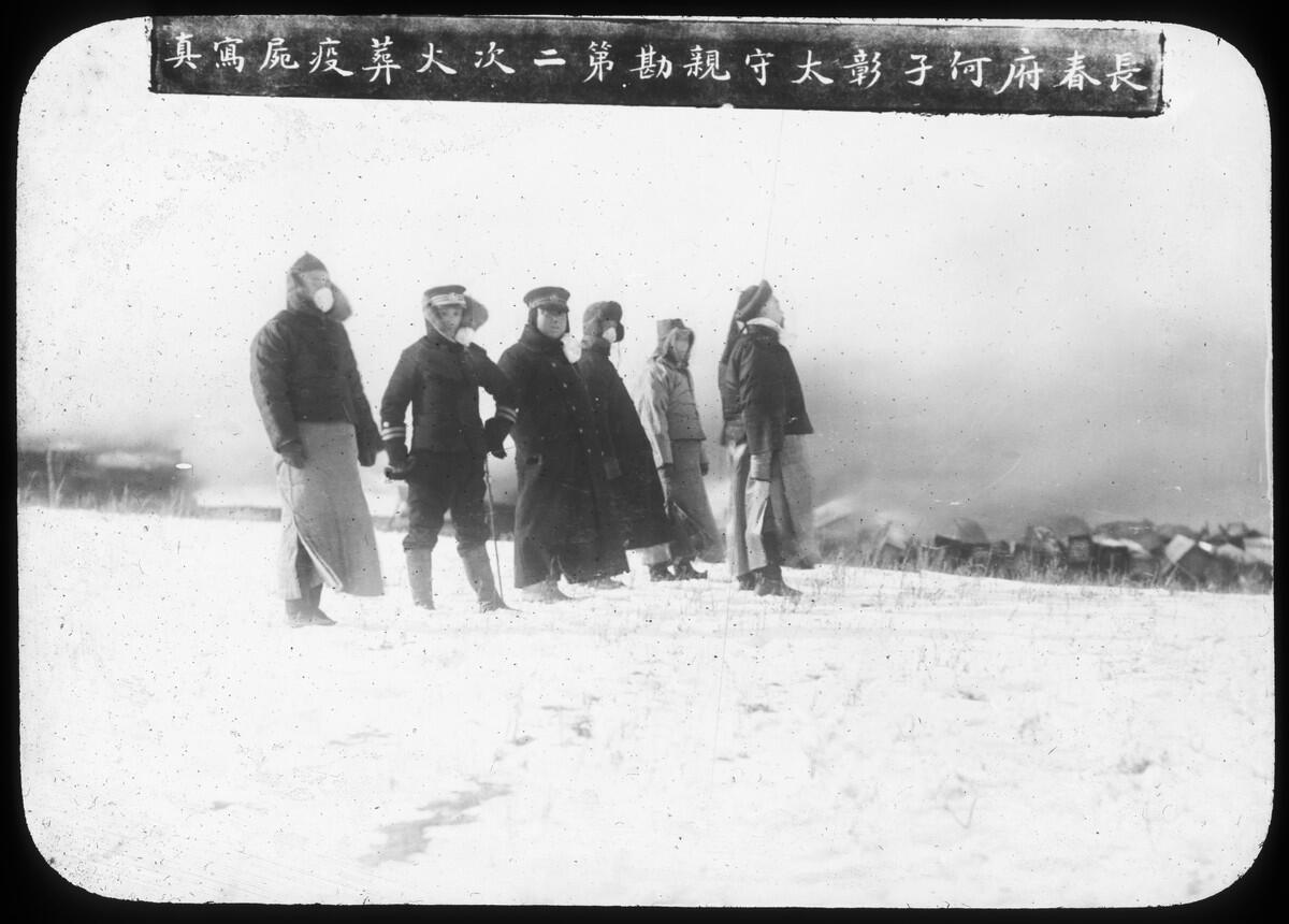 A group of men wearing protective gear and masks watch a cremation in the open air.