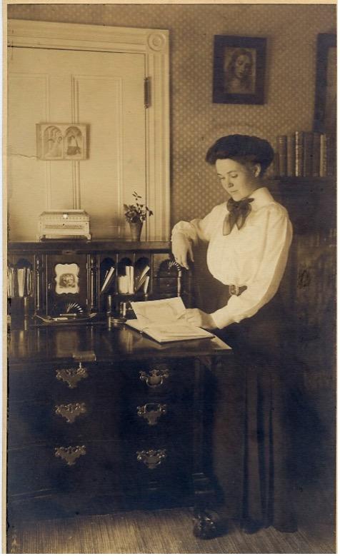 A young woman in long skirt looks down at a book upon a cabinet-style writing desk