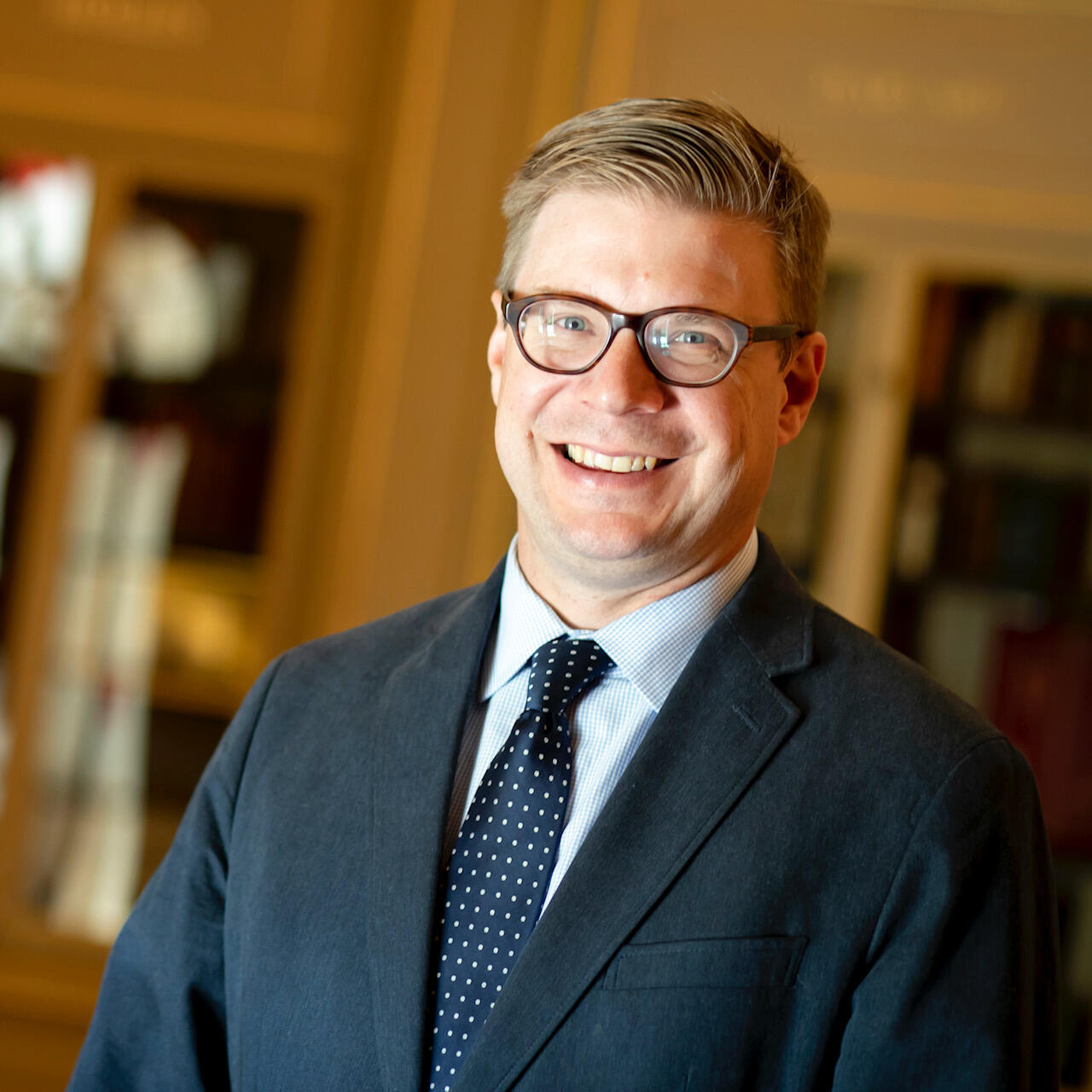 A white man wearing a suit, tie, and glasses smiles in front of glass-front bookcases.
