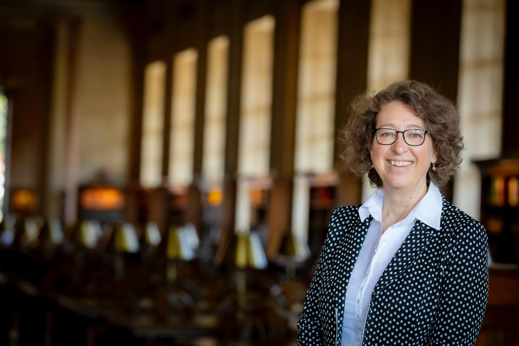 Portrait of Martha Whitehead wearing glasses, a black polka-dotted jacket, and white collared shirt, standing in the Loker Reading Room, with study tables and lamps in the background.