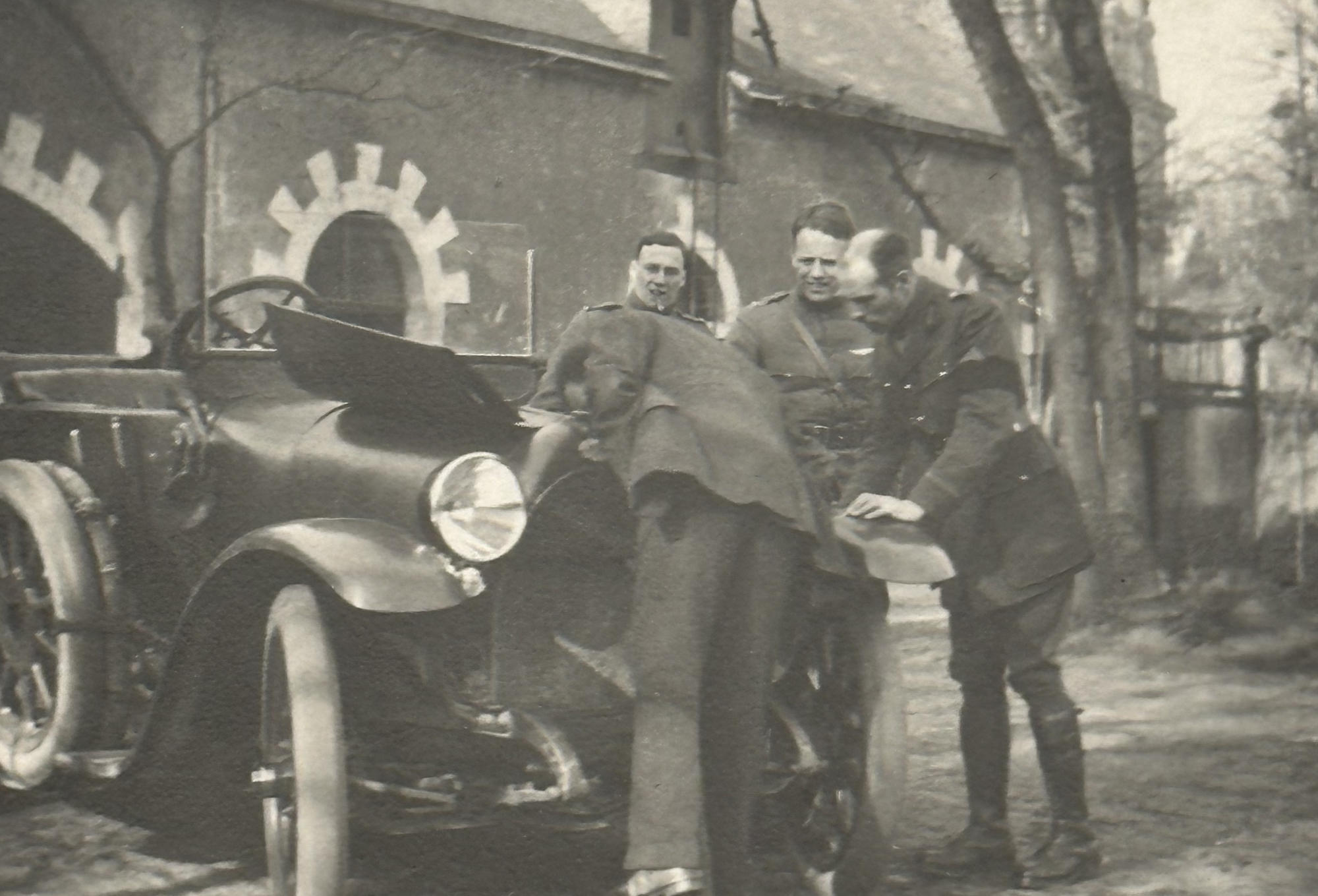 Four men stand around a car with the motor bonnet open in front of a large brick house