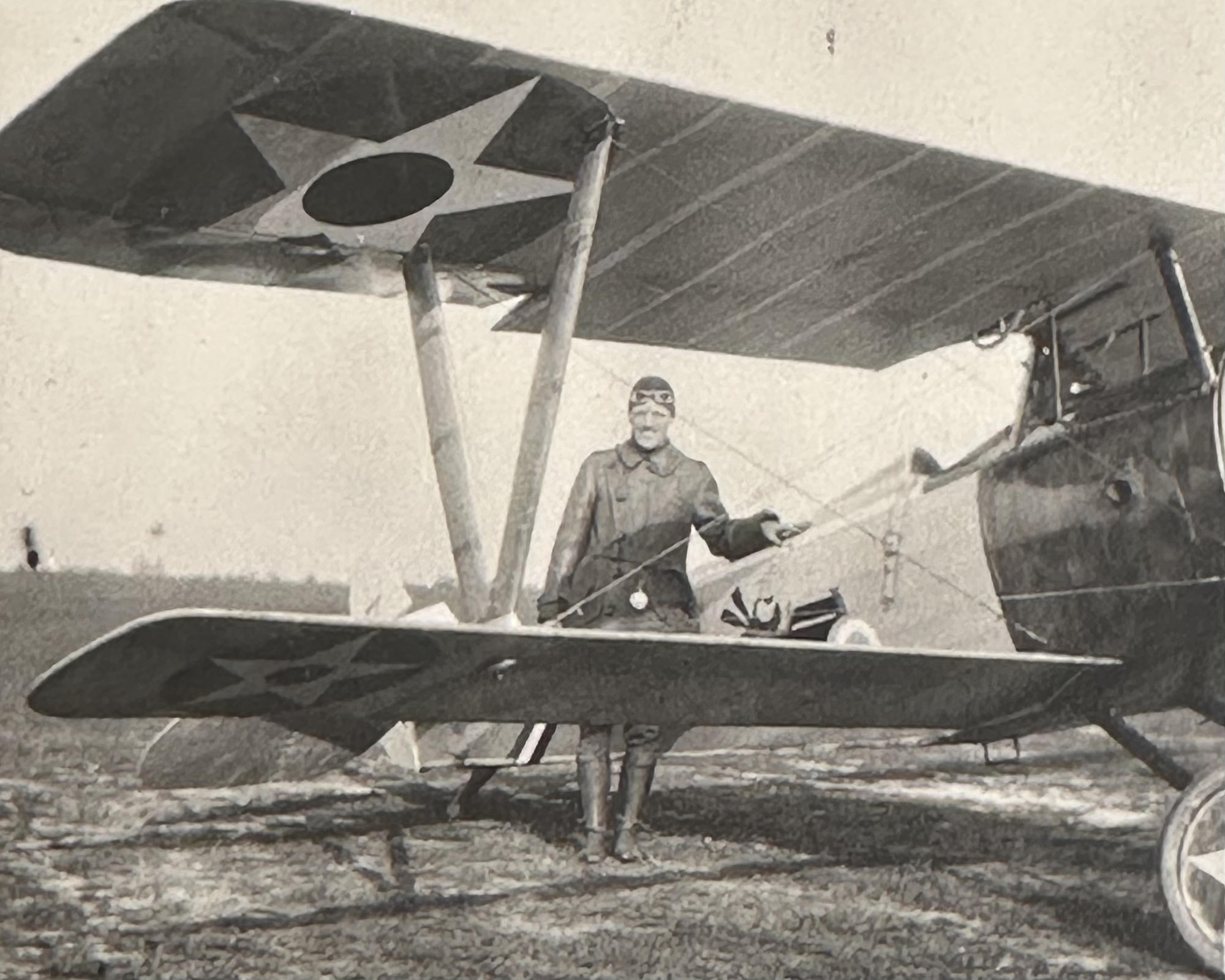 A man wearing a leather jacket, flight helmet and goggles stands next to a single engine fighter plane
