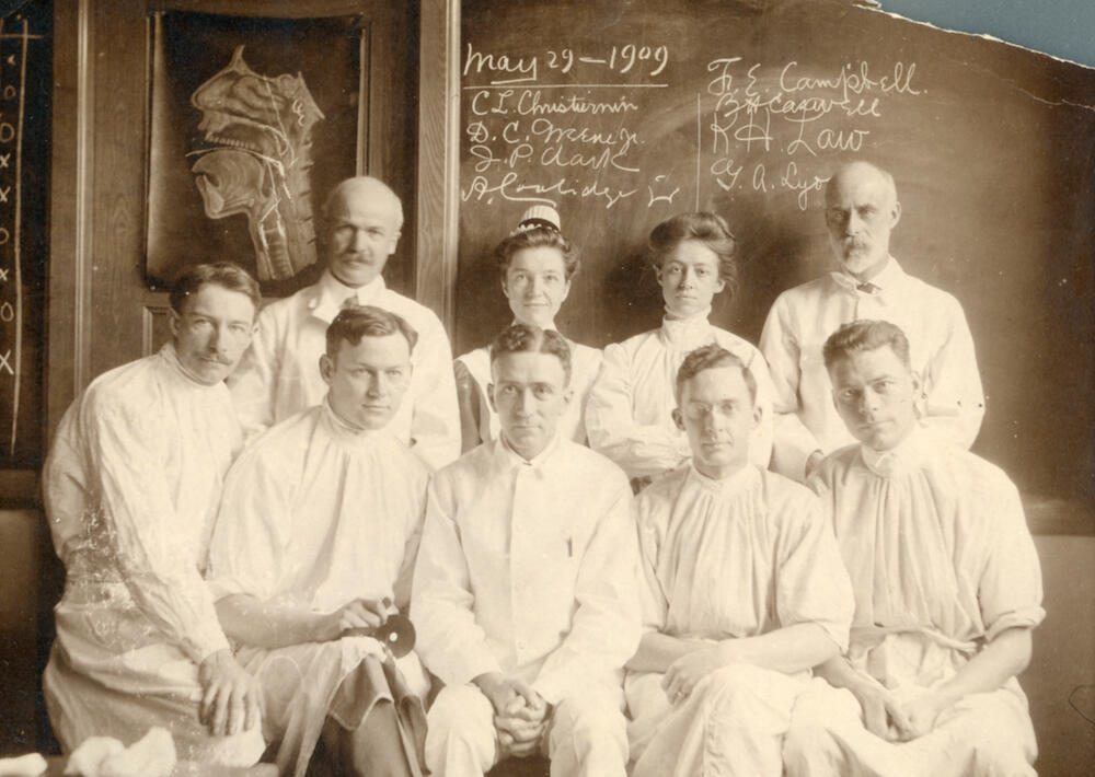 Nine white people, two women and seven men, pose in two rows wearing surgical gowns in front of a chalkboard and an anatomical illustration of the nose, mouth, and esophagus.