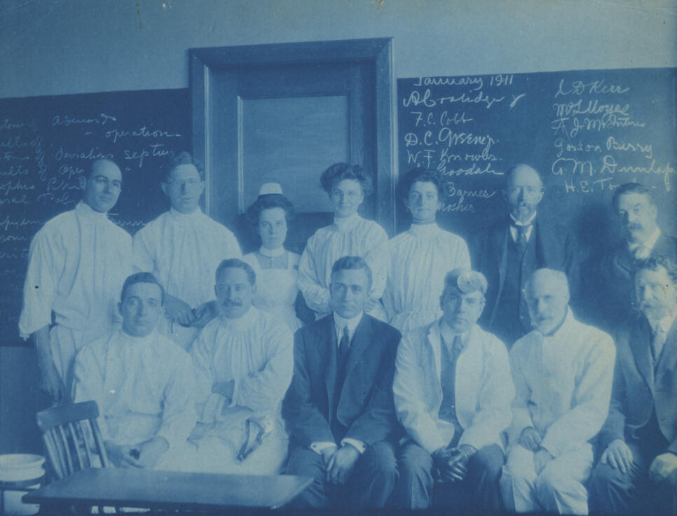 Thirteen white people, three women and ten men, pose in two rows with the front row sitting and back row standing. Behind them is a chalkboard and in front of them is a small table and chair.
